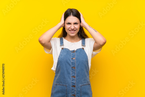 Young woman in dungarees over isolated yellow background frustrated and takes hands on head