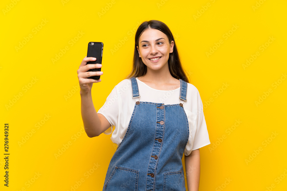 Young woman in dungarees over isolated yellow background making a selfie Young woman in dungarees over isolated yellow background making a selfie
