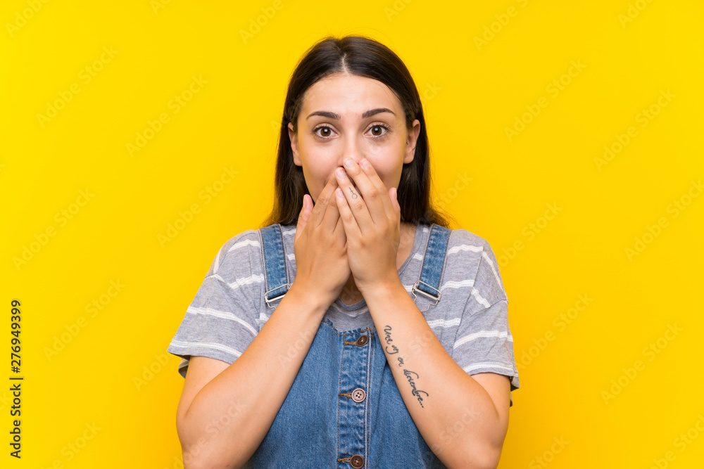 Young woman in dungarees over isolated yellow background with surprise facial expression Young woman in dungarees over isolated yellow background with surprise facial expression