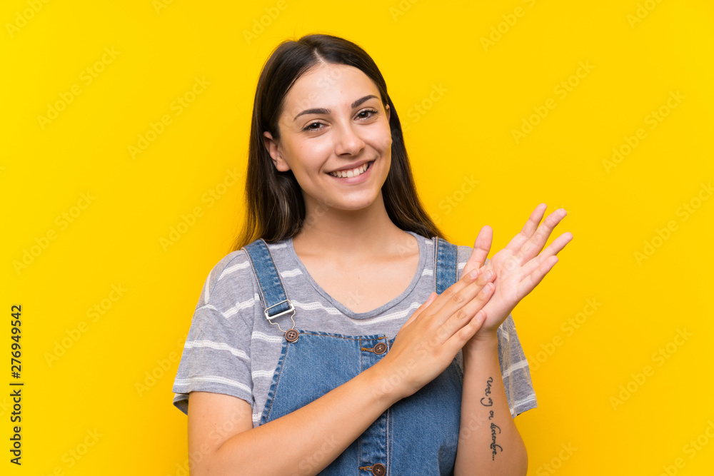 Young woman in dungarees over isolated yellow background applauding Young woman in dungarees over isolated yellow background applauding