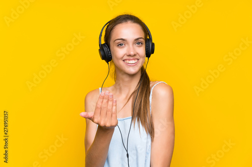Young woman listening music over isolated yellow wall inviting to come with hand. Happy that you came
