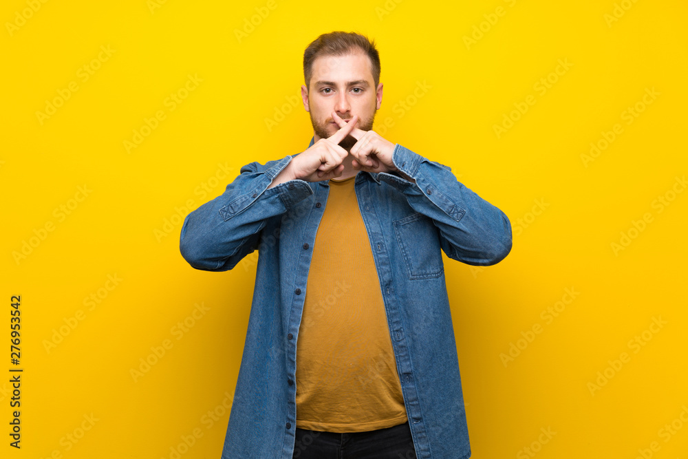 Blonde man over isolated yellow wall showing a sign of silence gesture Blonde man over isolated yellow wall showing a sign of silence gesture