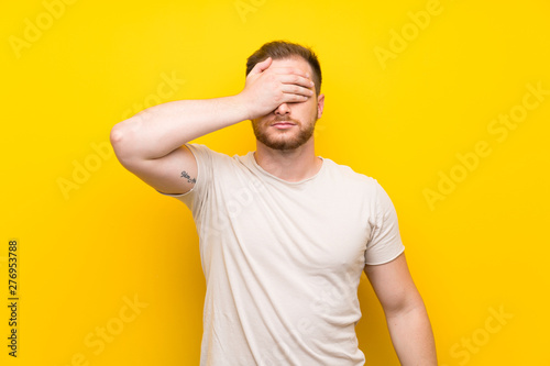 Handsome man over yellow background covering eyes by hands © luismolinero