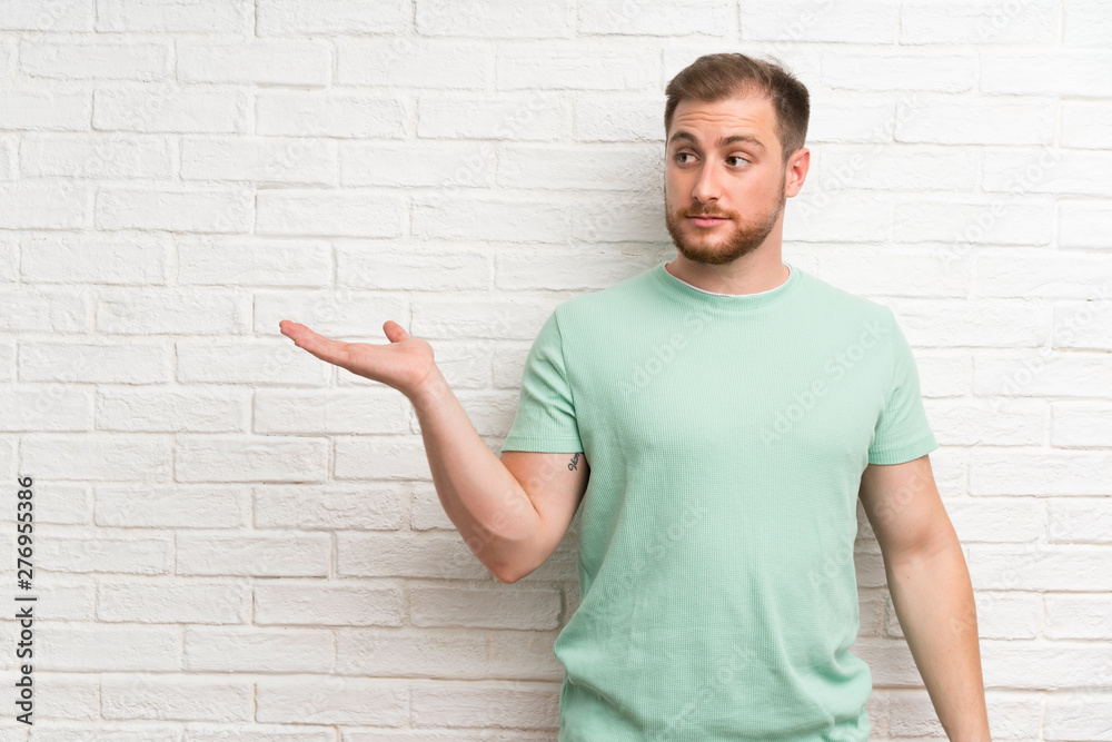 Blonde man over brick wall holding copyspace imaginary on the palm Blonde man over brick wall holding copyspace imaginary on the palm