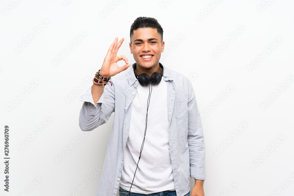 Young man over isolated white wall with earphones showing ok sign with fingers Young man over isolated white wall with earphones showing ok sign with fingers