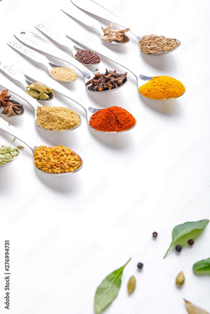 Various spices ( cumin, coriander, curry, paprika, chili, turmeric cinnamon, fenugreek, cardamom, basil leaf, parsley, cloves )  in  spoons on white background. Top view with copy space.