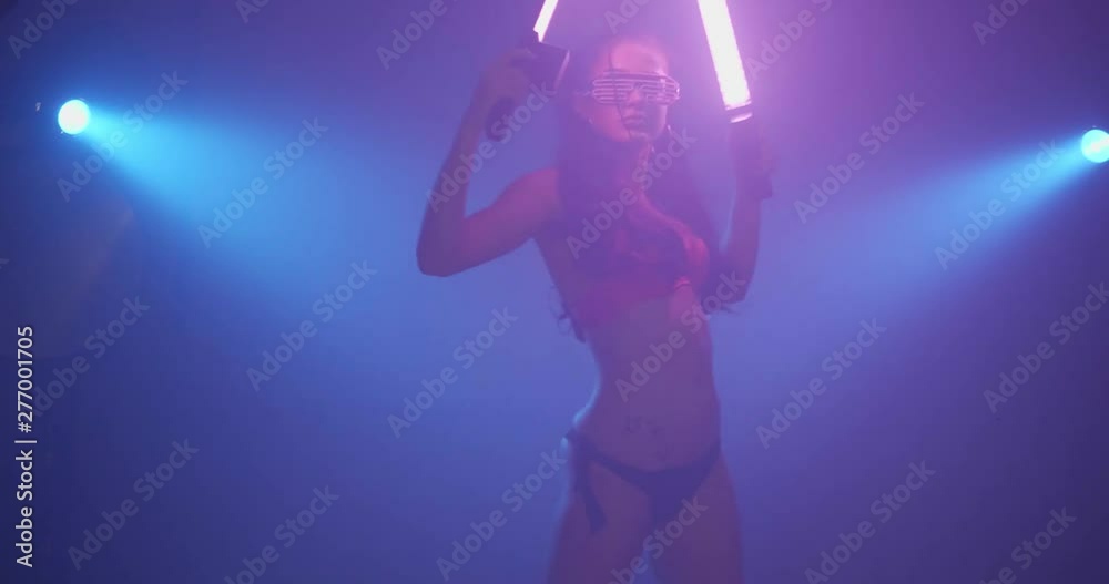 Party sensual woman wearing neon flashing pink bra and glasses dancing with glowing light sticks in hazy studio nightclub Party sensual woman wearing neon flashing pink bra and glasses dancing with glowing light sticks in hazy studio nightclub