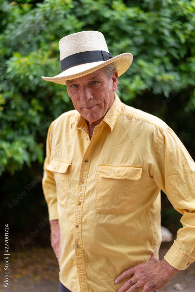 Portrait of good looking senior man in his 60s outdoors in backyard, wearing hat and yellow shirt.  Looking away from camera