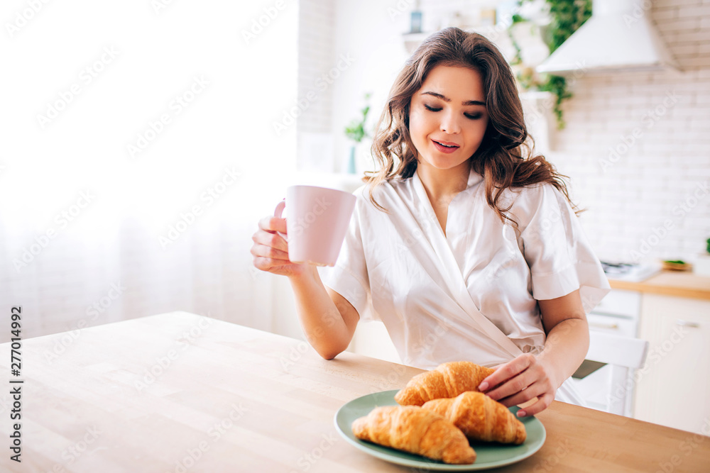 Young woman with dark hair Sitting in kitchen and drinking coffee in morning. Taking one croissant and smile. Delightful model. Young woman with dark hair Sitting in kitchen and drinking coffee in morning. Taking one croissant and smile. Delightful model.
