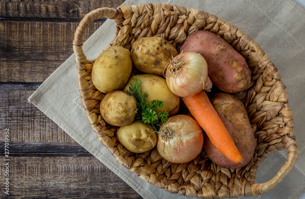 Wicker basket with vegetables on dark wooden background. Raw potatoes, onions, carrots and bunches green. Concept harvest. Flat lay composition. Wicker basket with vegetables on dark wooden background. Raw potatoes, onions, carrots and bunches green. Concept harvest. Flat lay composition.
