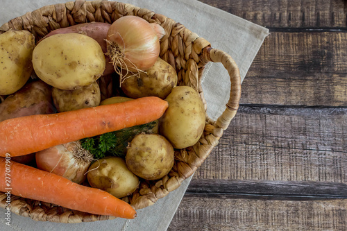 Wicker basket with vegetables on dark wooden background. Raw potatoes, onions, carrots and bunches green. Concept harvest. Flat lay composition. 