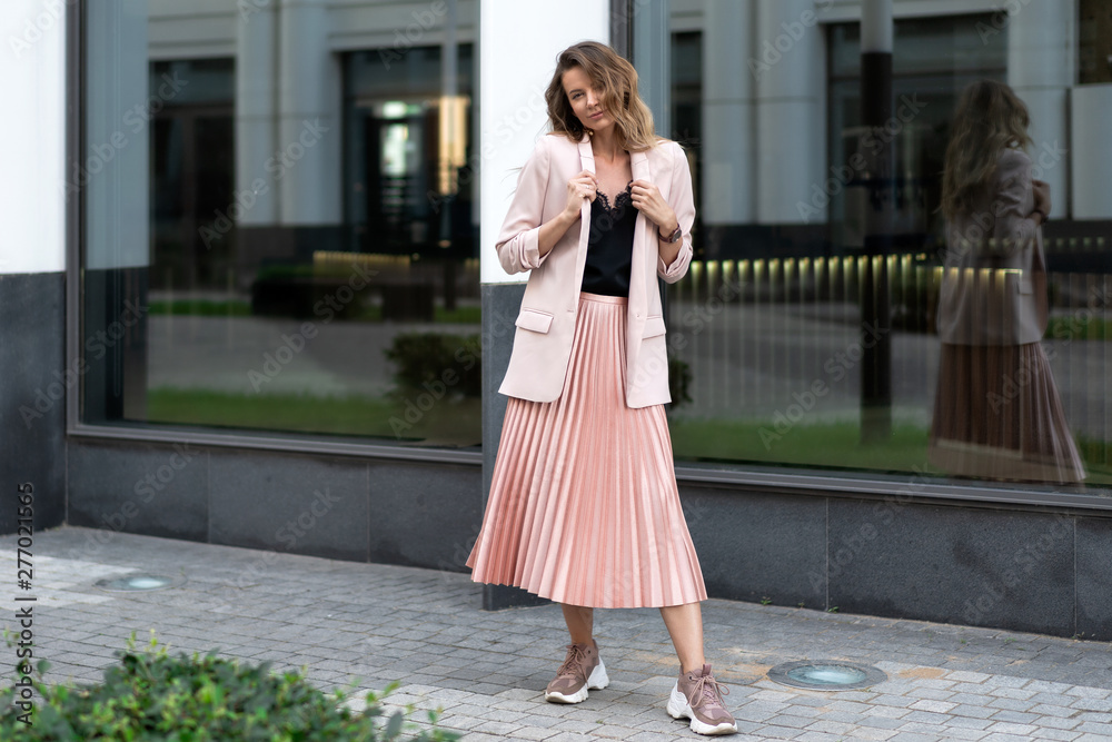 model posing in coral color clothes. Pleated long skirt, jacket, sneakers, black blouse. wavy hair below shoulders. Young beautiful european woman stands on the street near the glass wall.