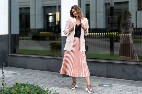 model posing in coral color clothes. Pleated long skirt, jacket, sneakers, black blouse. wavy hair below shoulders. Young beautiful european woman stands on the street near the glass wall.
