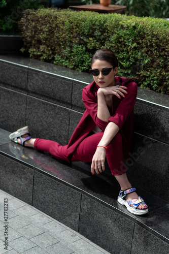 A young girl poses in an original pose in a burgundy business trouser suit. Women's jacket and pants in red. Model with a short haircut.