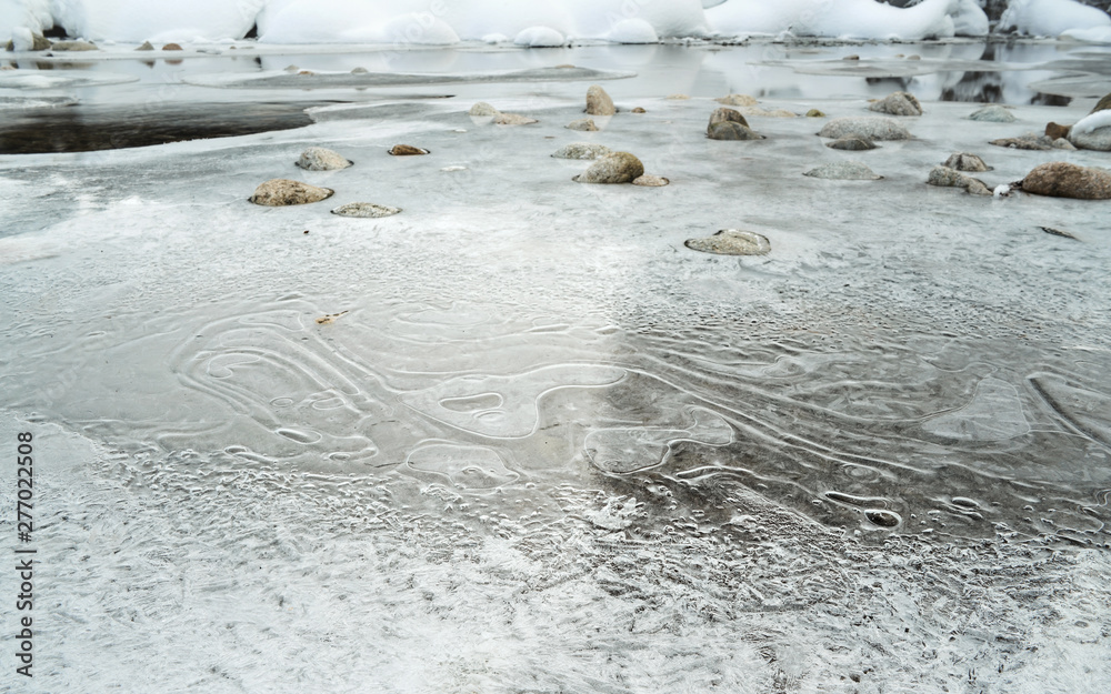 Φωτογραφία Closeup of thin ice and crystalline snow layer on frozen river in winter, low angle photo, elliptical ice patterns and water flowing under, stones, in distance