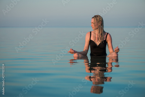 young woman in swim suit sitting in yoga pose in calm sea water at summer sunset