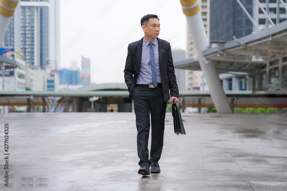 Asian Businessman walking and holding briefcase with business office buildings in the city background