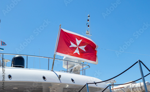 Maltese old flag on pole on ferry's stern. Ongoing cruise to islands. Blue sky background, close up view.