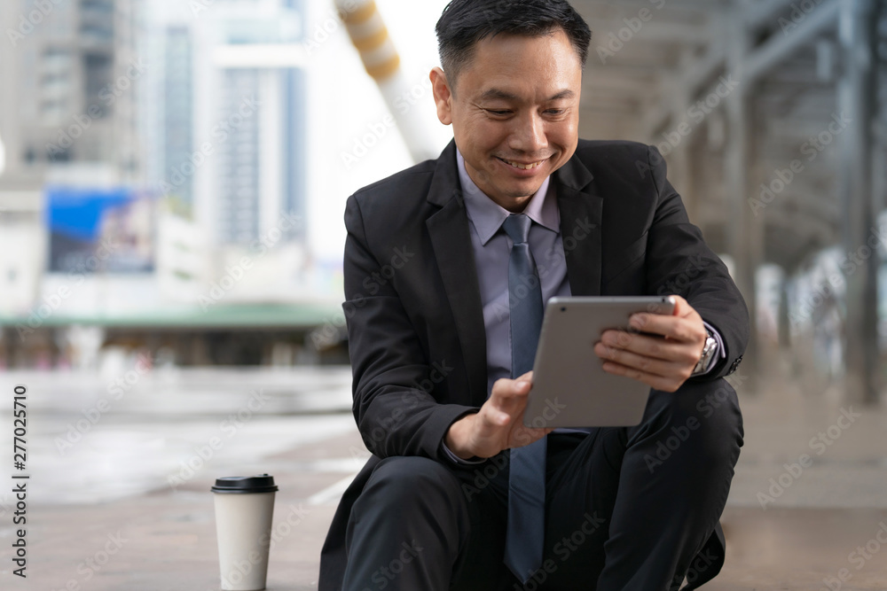 Asian Businessman sitting and holding digital tablet with business office buildings in the city background Asian Businessman sitting and holding digital tablet with business office buildings in the city background