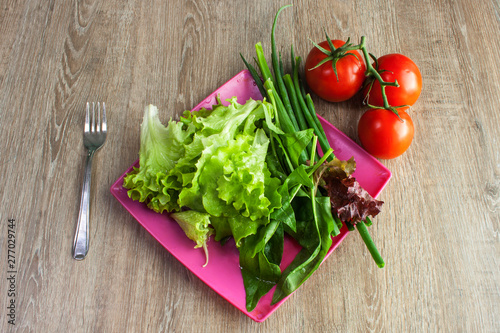 Salad with greens, cherry tomatoes, onions on pink square plate with fork on wooden table, overhead view
