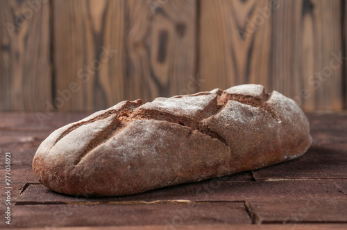 Whole loaf of dark homemade bread dusted with flour is lying on old brown boards