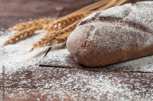 Whole loaf of dark homemade bread sprinkled with flour near bunch of wheat lies on old brown boards
