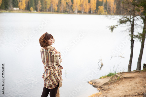 Golden autumn shooting on the lake girl stands back, brunette with curls, wrapped in a warm blanket of light brown. the scarf is knit.