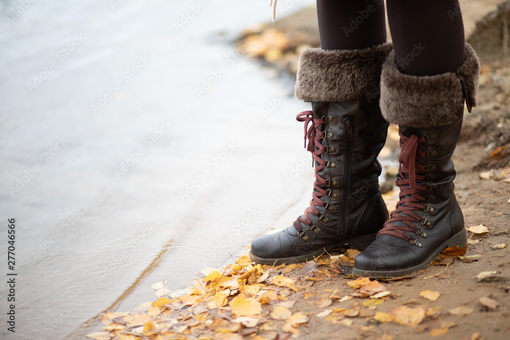 A girl in brown boots stands on the sandy shore of the lake around the yellow leaves, water, close-up, boots with fur on the laces to the knee.