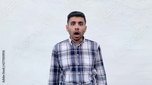 Portrait of shocked handsome bearded man in blue checkered shirt standing and looking at camera with big eyes and unbelievable face. indoor studio shot isolated on white wall background.