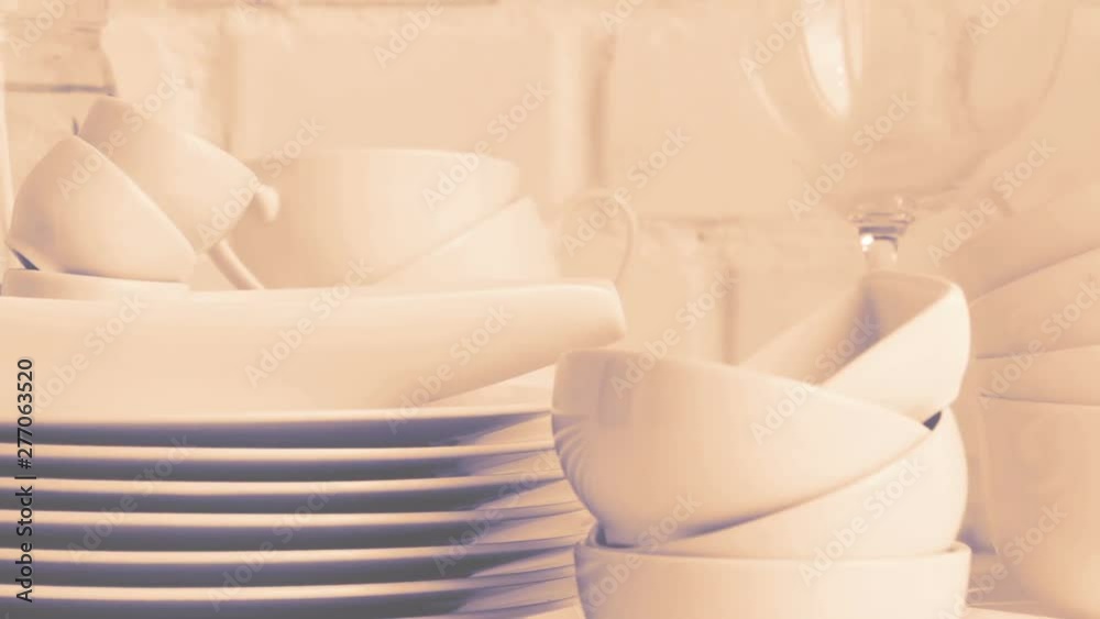 Foggy and warm toned. Stacked white porcelain dishes, plates, cups, saucers, mugs in restaurant kitchen interior. Slow pan. White brick wall background. Selective focus. Foggy and warm toned. Stacked white porcelain dishes, plates, cups, saucers, mugs in restaurant kitchen interior. Slow pan. White brick wall background. Selective focus.