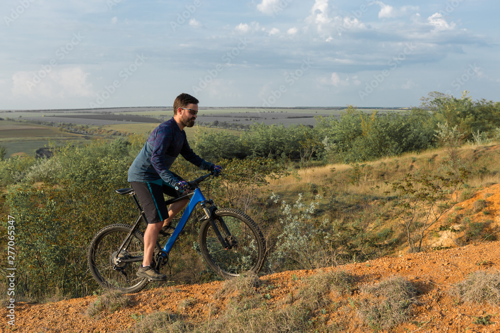 Cyclist in shorts and jersey on a modern carbon hardtail bike with an air suspension fork rides off-road on the orange-red hills at sunset evening in summer Cyclist in shorts and jersey on a modern carbon hardtail bike with an air suspension fork rides off-road on the orange-red hills at sunset evening in summer
