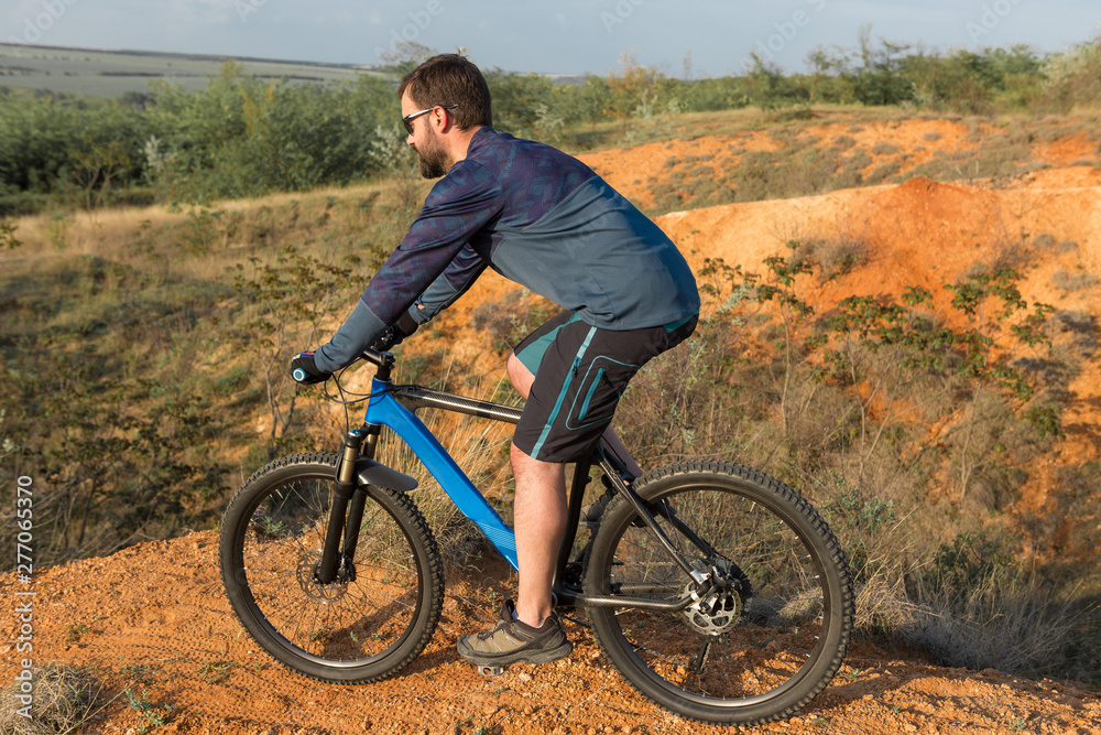 Fototapeta premium Cyclist in shorts and jersey on a modern carbon hardtail bike with an air suspension fork rides off-road on the orange-red hills at sunset evening in summer 