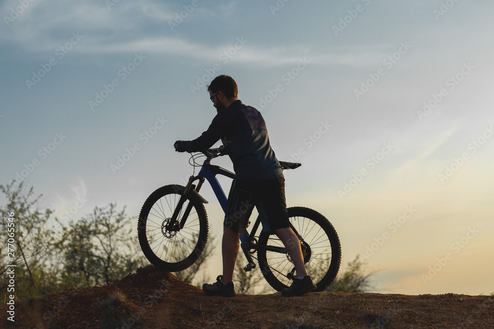 Cyclist in shorts and jersey on a modern carbon hardtail bike with an air suspension fork rides off-road on the orange-red hills at sunset evening in summer Cyclist in shorts and jersey on a modern carbon hardtail bike with an air suspension fork rides off-road on the orange-red hills at sunset evening in summer