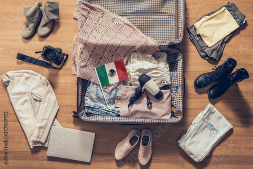 Woman's clothes, laptop, camera and flag of Mexico lying on the parquet floor near and in the open suitcase. Travel concept