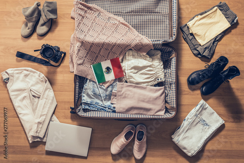 Woman's clothes, laptop, camera and flag of Mexico lying on the parquet floor near and in the open suitcase. Travel concept