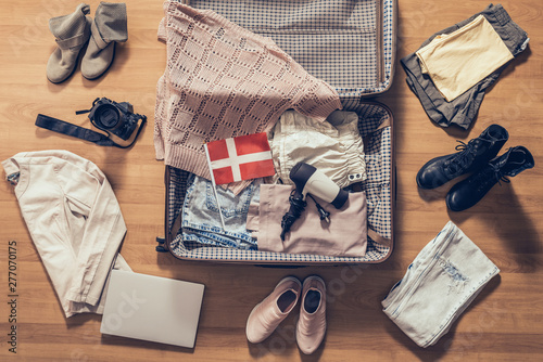 Woman's clothes, laptop, camera and flag of Denmark lying on the parquet floor near and in the open suitcase. Travel concept