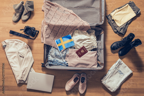 Woman's clothes, laptop, camera, russian passport and flag of Sweden lying on the parquet floor near and in the open suitcase. Travel concept