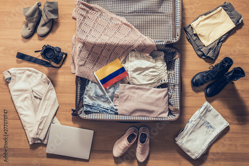 Woman's clothes, laptop, camera and flag of Colombia lying on the parquet floor near and in the open suitcase. Travel concept