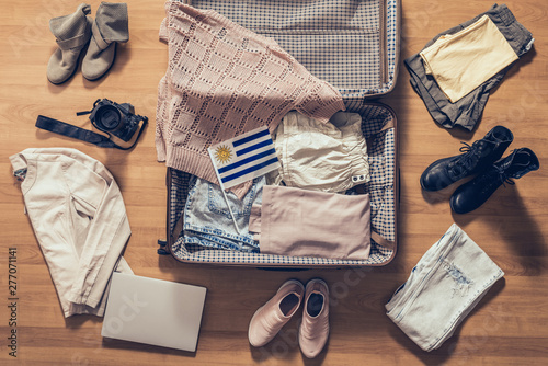 Woman's clothes, laptop, camera and flag of Uruguay lying on the parquet floor near and in the open suitcase. Travel concept