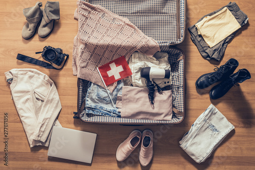 Woman's clothes, laptop, camera and flag of Switzerland lying on the parquet floor near and in the open suitcase. Travel concept