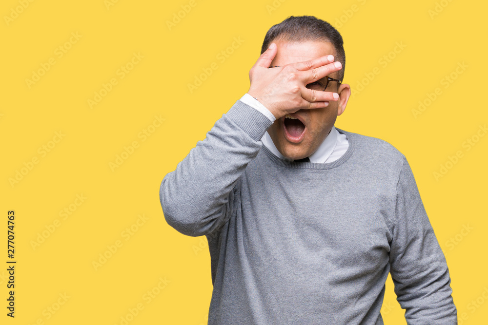 Middle age bussines arab man wearing glasses over isolated background peeking in shock covering face and eyes with hand, looking through fingers with embarrassed expression. Middle age bussines arab man wearing glasses over isolated background peeking in shock covering face and eyes with hand, looking through fingers with embarrassed expression.