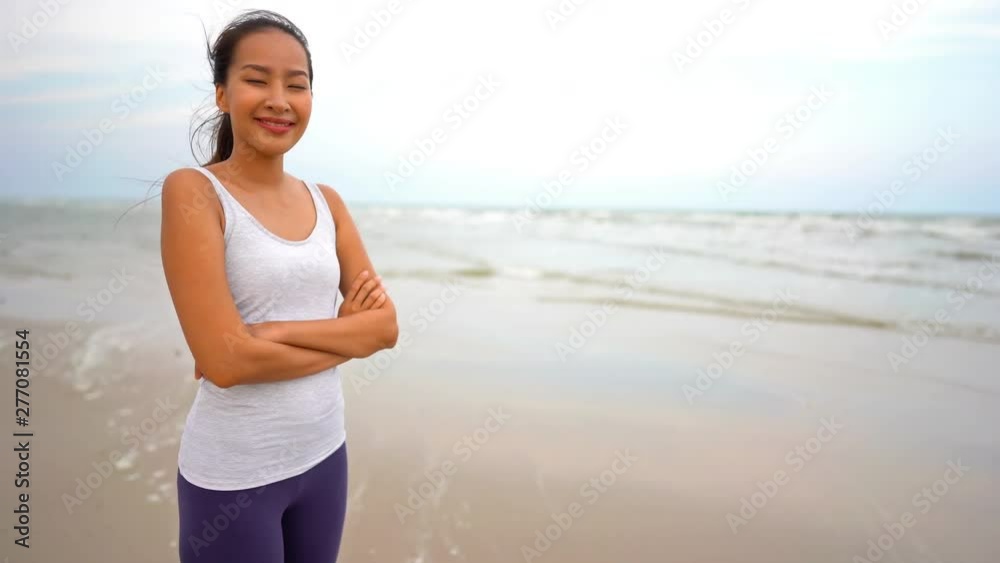 Bust shot, asian young lady smiling to the camera at the beach in front of the sea wearing white sports jersey and grey leggings Bust shot, asian young lady smiling to the camera at the beach in front of the sea wearing white sports jersey and grey leggings