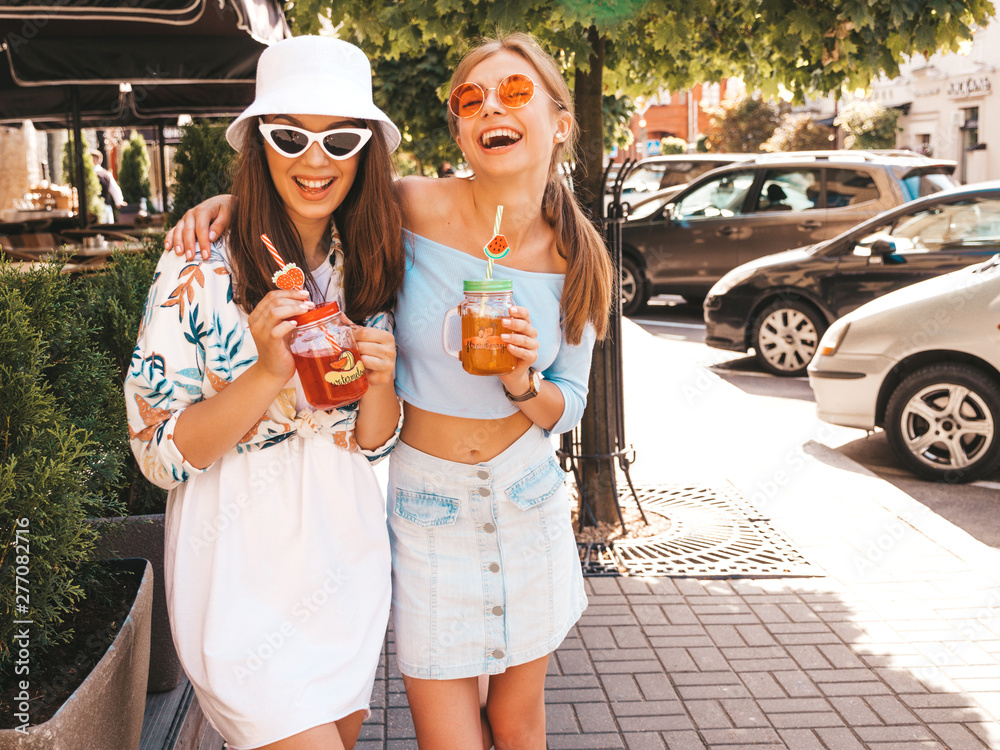 Fototapeta premium Two young beautiful smiling hipster girls in trendy summer clothes and panama hat.Sexy carefree women posing in the street.Positive models having fun in sunglasses.Drinking fresh cocktail smoozy drink
