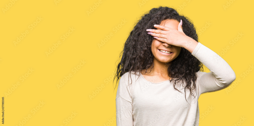 Young beautiful girl with curly hair wearing casual sweater smiling and laughing with hand on face covering eyes for surprise. Blind concept. Young beautiful girl with curly hair wearing casual sweater smiling and laughing with hand on face covering eyes for surprise. Blind concept.