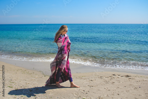 Blonde girl have fun on the beach. Young smiling woman wrapped in warm pink blanket near sea.