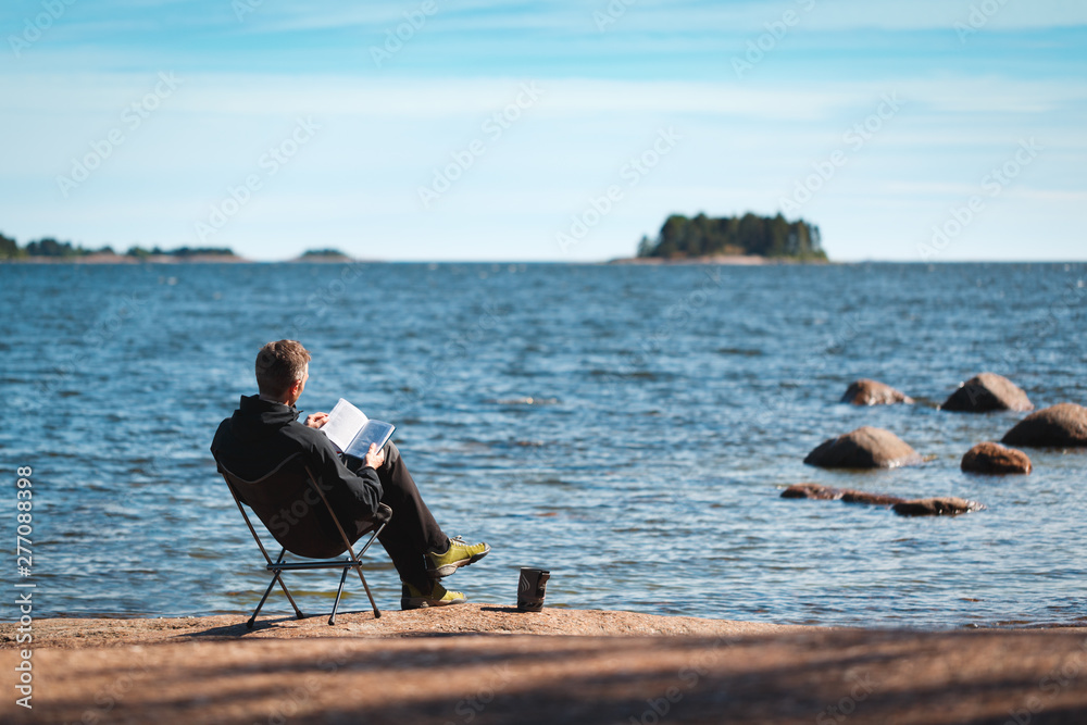 A man reading a book on the beach sitting on a chair. Near mug. Object in focus , background blurred