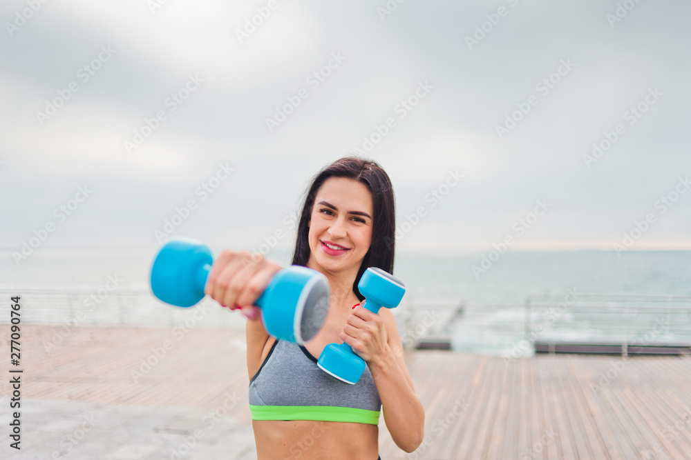 Young cheerful fit woman trains straight arm punch with dumbbells on the beach at sunrise Young cheerful fit woman trains straight arm punch with dumbbells on the beach at sunrise