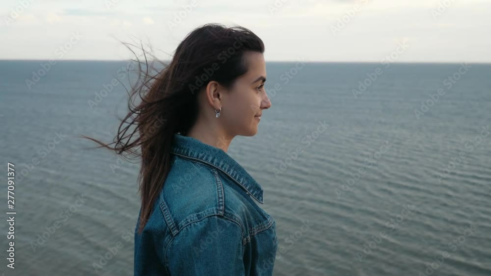 Portrait of young beautiful brunette lady standing on the mountain near the sea with strong wing waving her hair at sunset in slow motion Portrait of young beautiful brunette lady standing on the mountain near the sea with strong wing waving her hair at sunset in slow motion