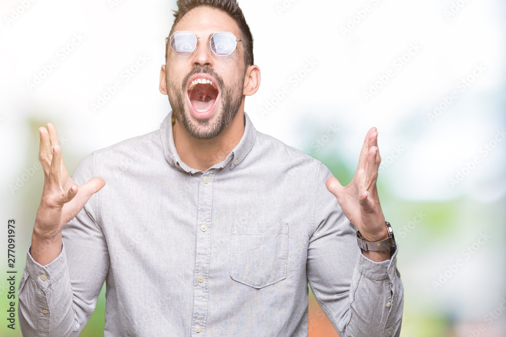 Naklejka premium Young handsome man wearing sunglasses over isolated background crazy and mad shouting and yelling with aggressive expression and arms raised. Frustration concept.