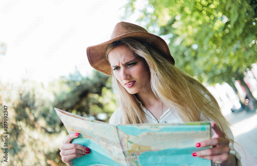 Blonde woman in a felt hat looks at city map and walks through an unfamiliar city outdoor